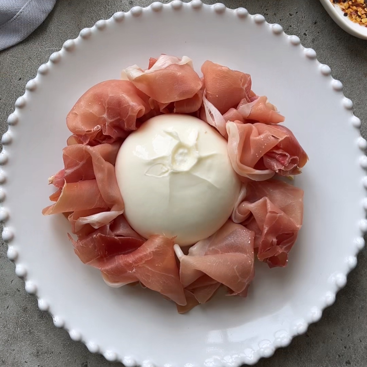 A ball of burrata cheese surrounded by folded slices of prosciutto on a white plate with a beaded rim.