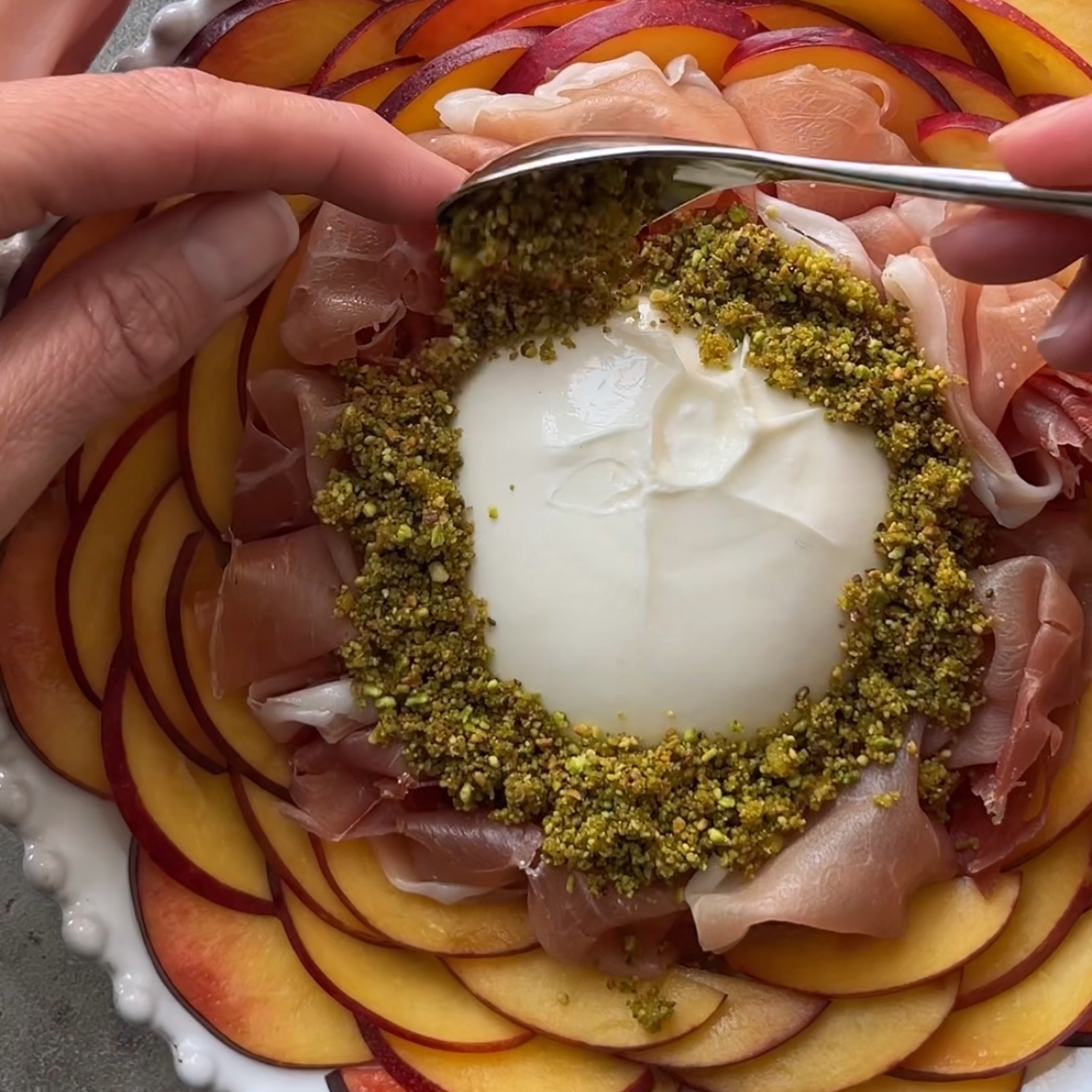 A hand sprinkles a crushed pistachio mixture onto burrata cheese, surrounded by prosciutto and a ring of sliced nectarines on a white plate.