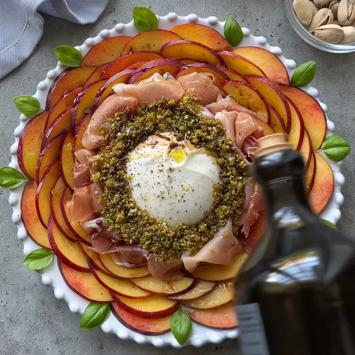 A plate with overlapping nectarine slices, prosciutto, burrata, crushed pistachios, and basil leaves resembles a refreshing Nectarine Salad. A hand pours olive oil over the center, while a bowl of pistachios sits in the background.
