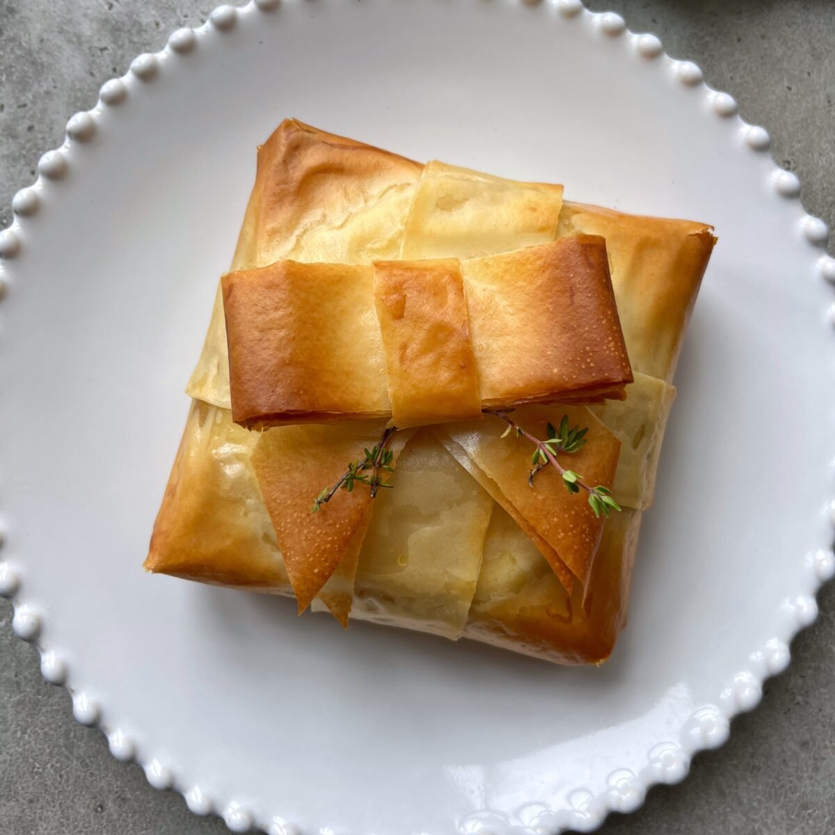A golden-brown phyllo baked brie resembling a gift, topped with delicate pastry strips and garnished with sprigs of fresh thyme, served on a white plate.