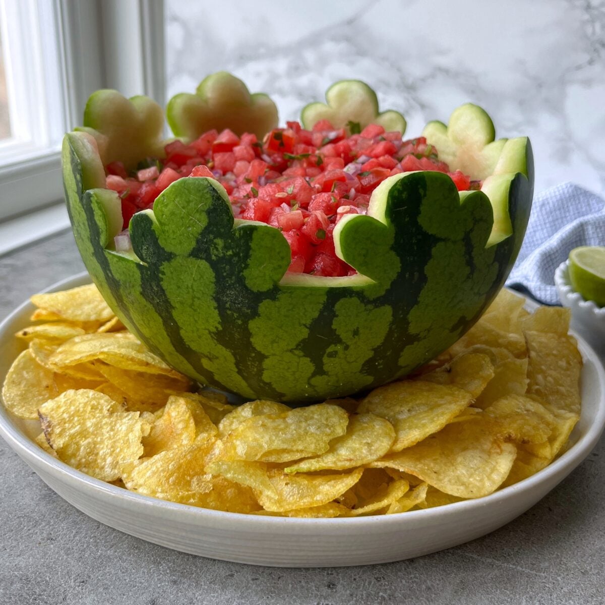 A carved watermelon bowl filled with Watermelon Salsa sits on a plate surrounded by potato chips on a gray countertop.