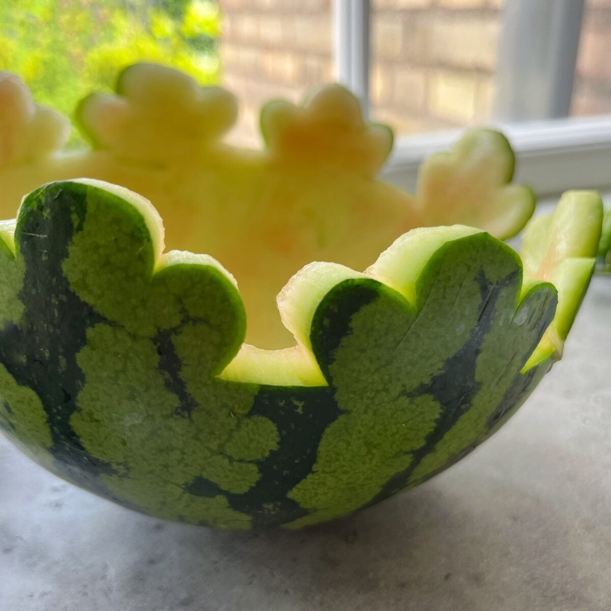 A hollowed watermelon half with a decorative scalloped edge sits on a countertop near a window, ready to be filled with refreshing Watermelon Salsa.