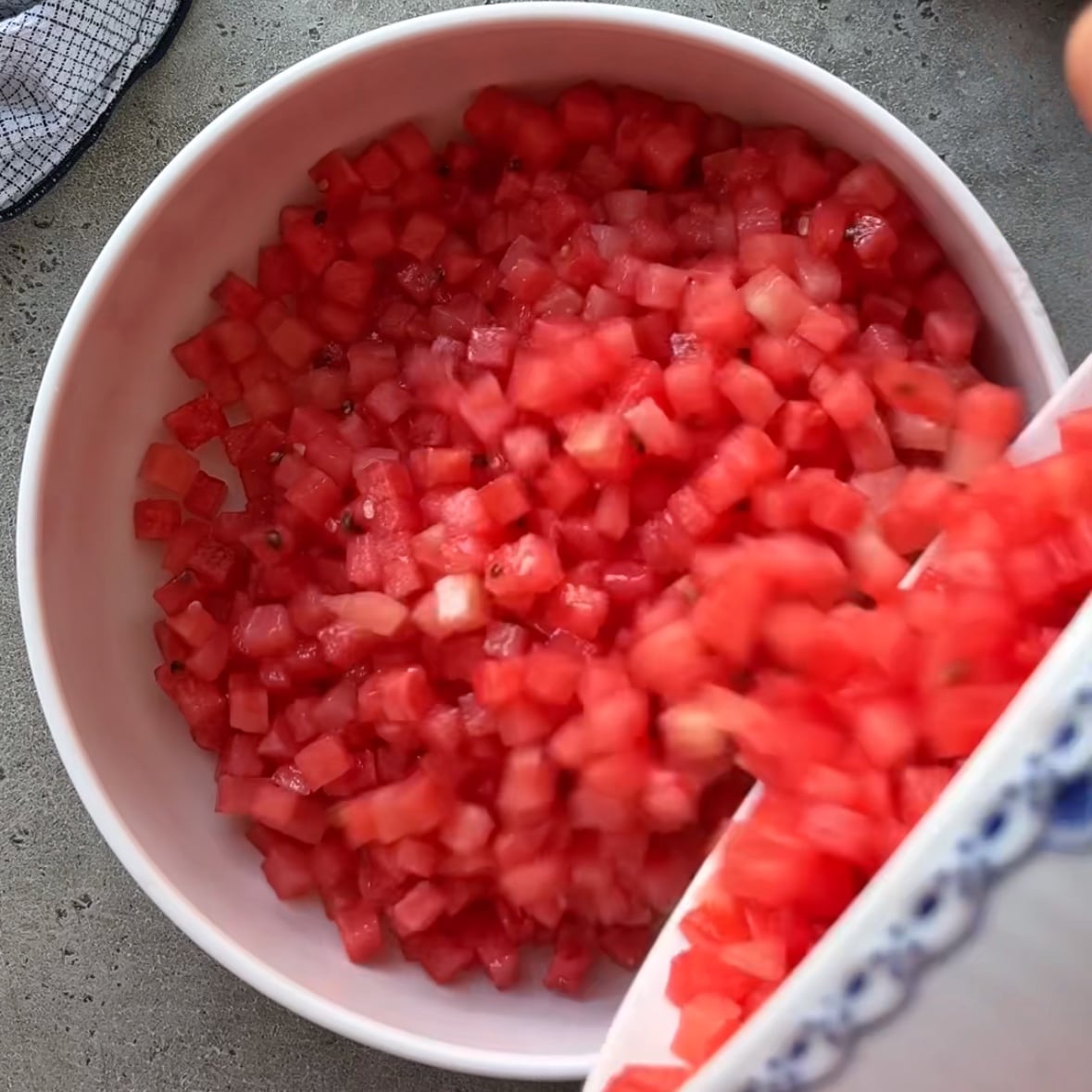Diced watermelon pieces are being poured from one bowl into a larger white bowl on a gray countertop, ready to be transformed into a refreshing Watermelon Salsa.