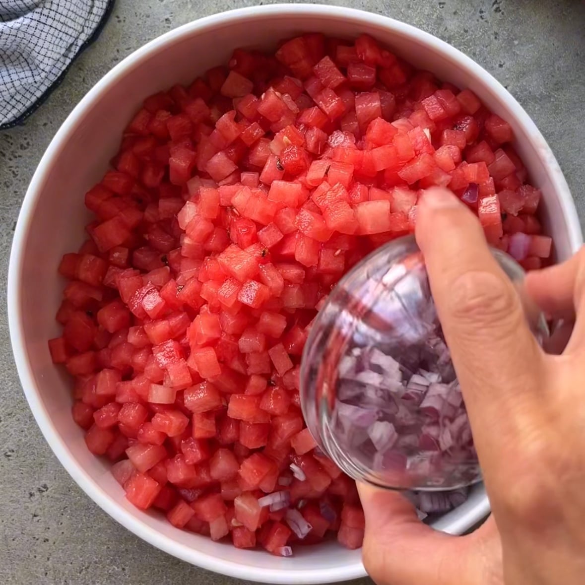 A hand adds diced red onions from a small glass bowl to a white bowl filled with chopped watermelon on a gray countertop.