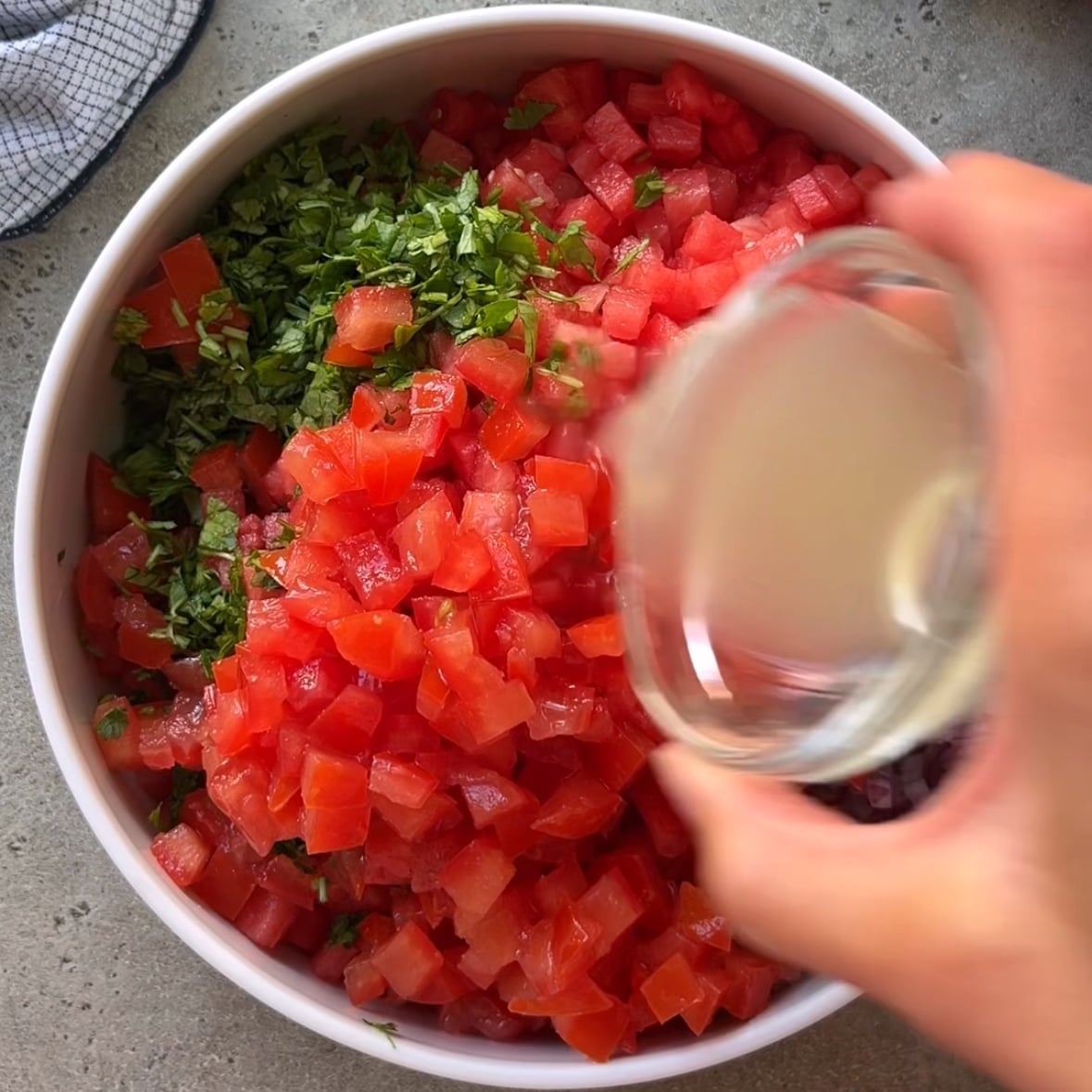 A hand pours a citrusy dressing into a bowl of diced watermelon, tomatoes, and chopped cilantro on a gray surface, preparing a fresh batch of watermelon salsa.