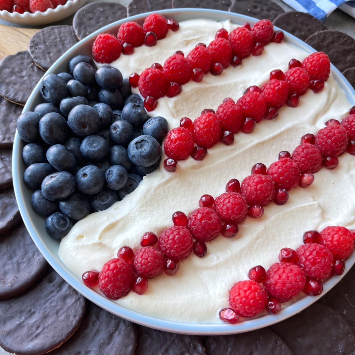 A dish decorated like an American flag with blueberries, white cream, raspberries, and pomegranate seeds, surrounded by chocolate cookies.