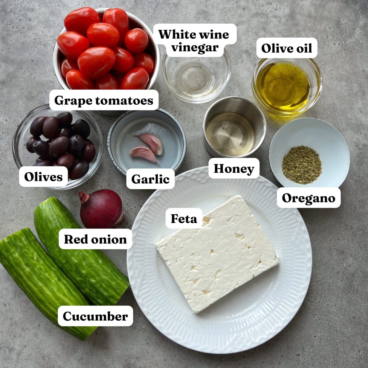 Ingredients for a Cucumber Tomato Feta Salad featuring grape tomatoes, olives, garlic, cucumber, red onion, feta, white wine vinegar, olive oil, honey, and oregano arranged on a table.