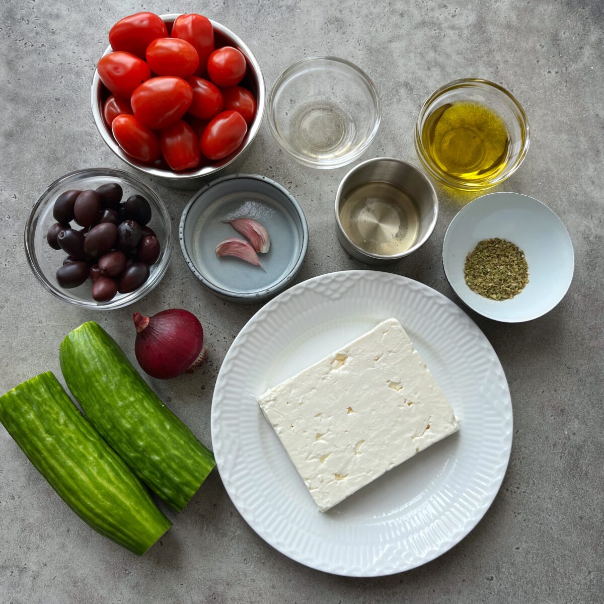 A plate of feta cheese surrounded by bowls of cherry tomatoes, kalamata olives, olive oil, vinegar, oregano, garlic, red onion, and cucumbers.