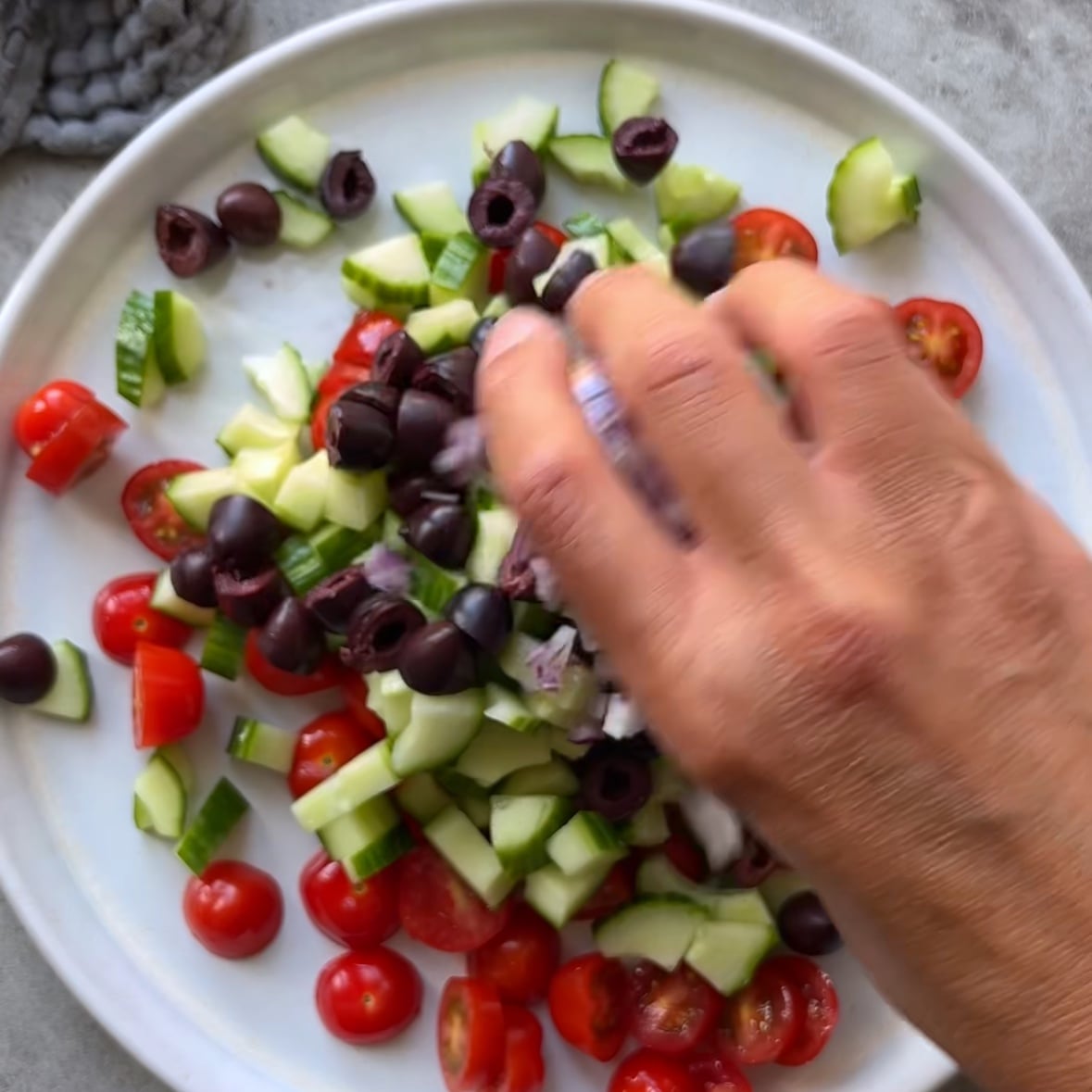 A hand adds red onion to other chopped ingredients on a white platter including cucumber, cherry tomatoes, and black olives.