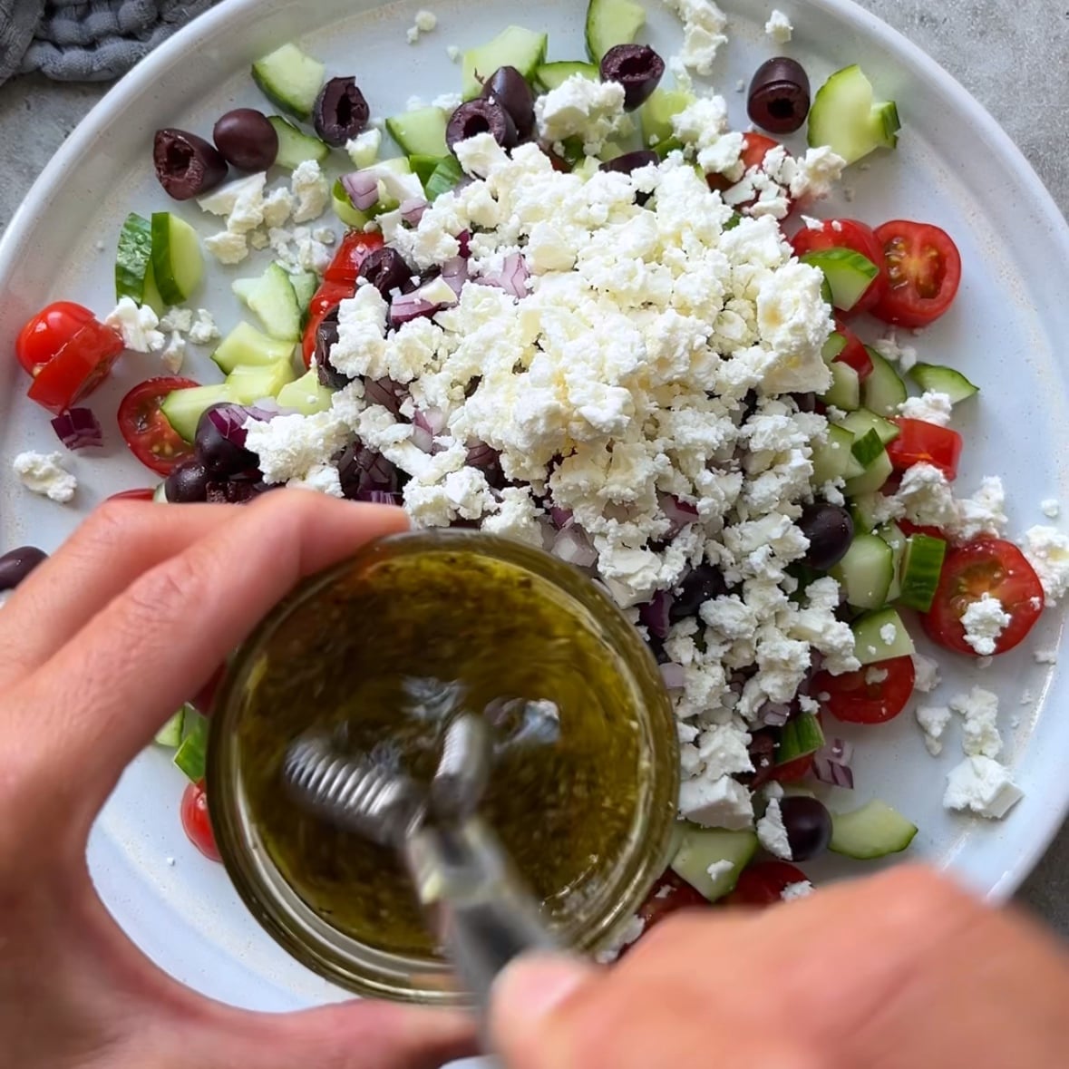A hand whisks dressing in a glass bowl above a plate of vibrant Cucumber Tomato Feta Salad, brimming with fresh cherry tomatoes, crisp cucumber, olives, and red onion.