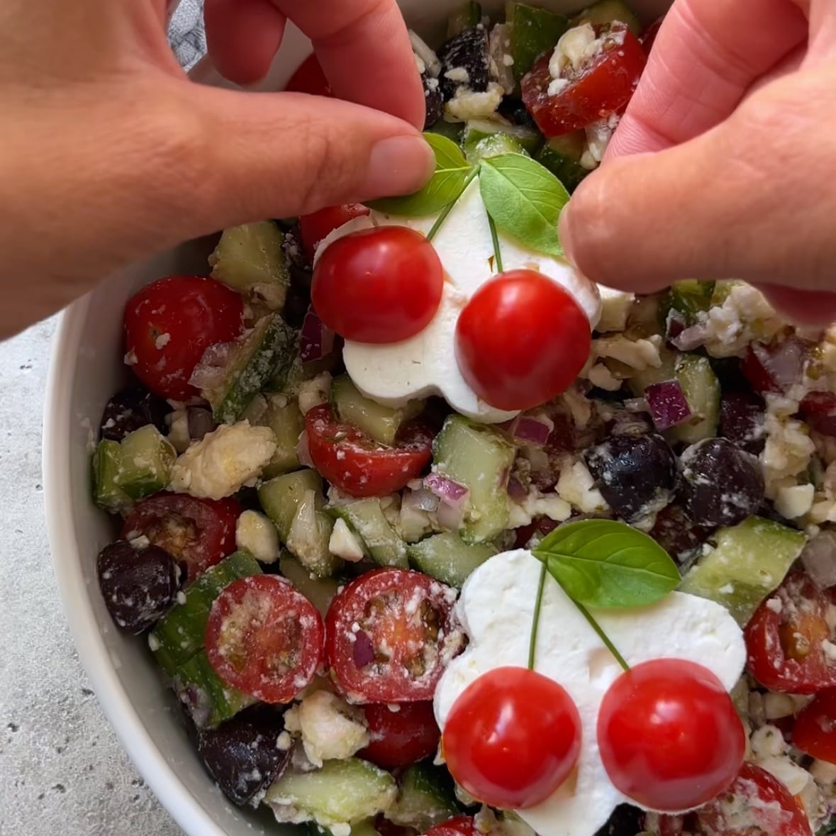 Hands hold cherry tomatoes and basil leaves shaped like a cherries over a vibrant Cucumber Tomato Feta Salad, featuring cucumbers, tomatoes, olives, feta, and onions in a white bowl.