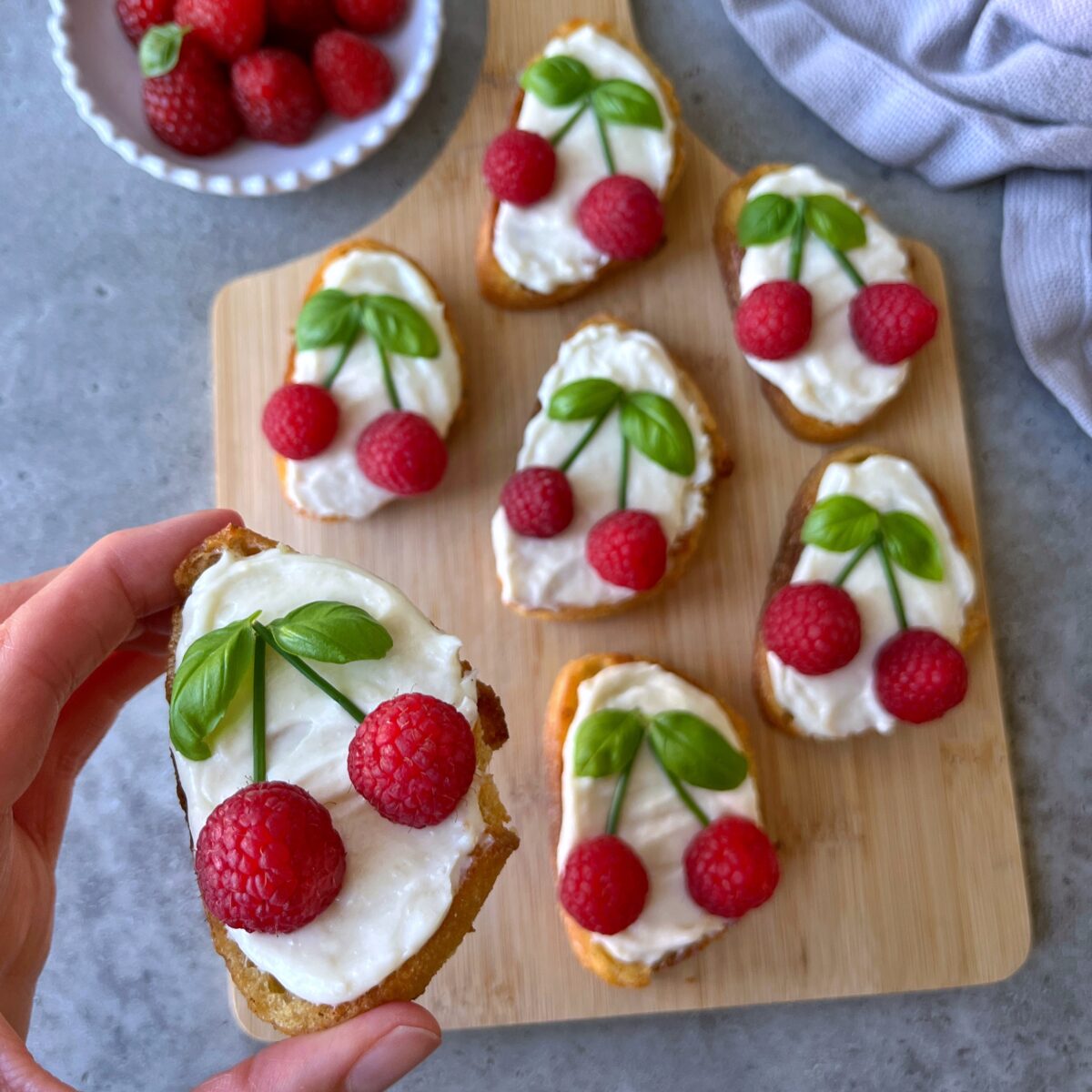 A hand holds a goat cheese crostini topped with two raspberries, and basil leaves to resemble a cherry.