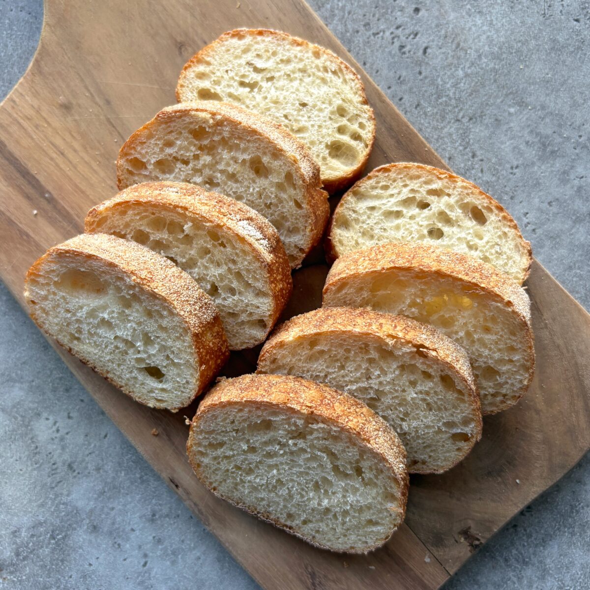 Eight slices of rustic bread are arranged in a row on a wooden cutting board, set on a gray textured surface.