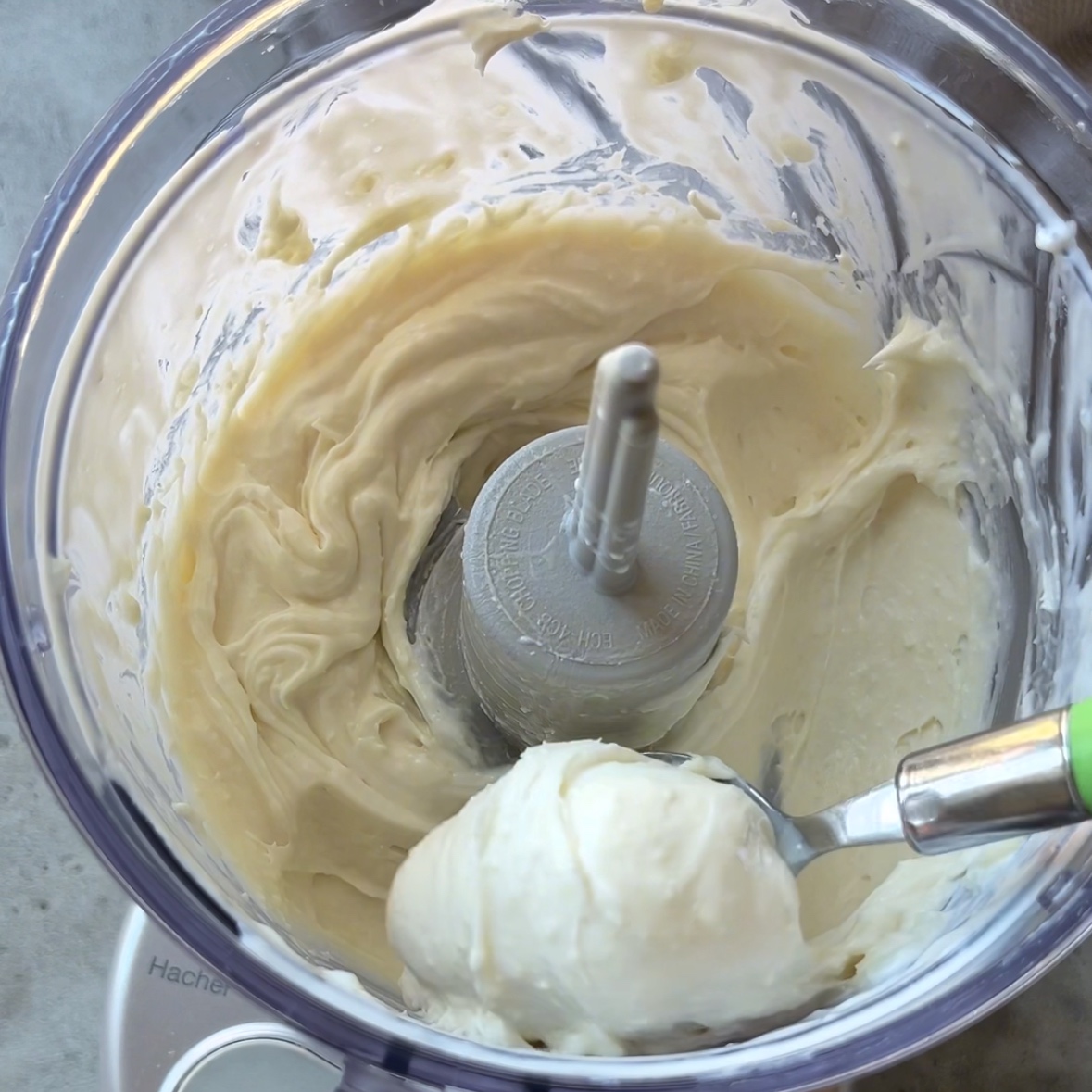 A creamy, pale goat cheese mixture is being scooped from a food processor with a spoon.