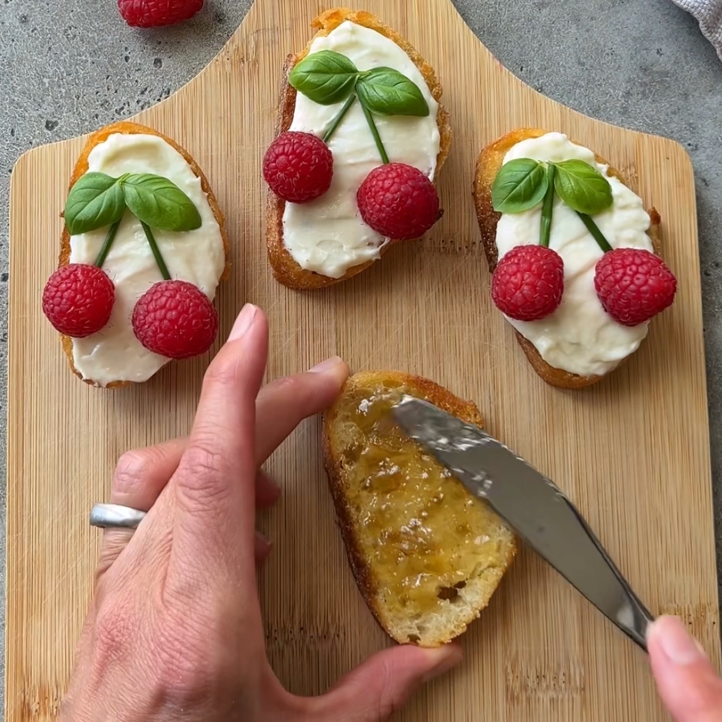A hand spreads jam on bread next to three Goat Cheese Crostini pieces topped with cream, raspberries, and basil leaves arranged to resemble cherries on a wooden board.