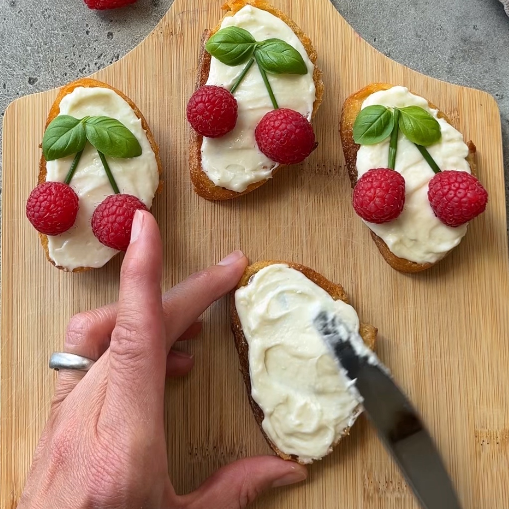 A hand spreads cream cheese on toast while three other toasts are topped with cream, raspberries, and basil leaves and arranged on a wooden board.