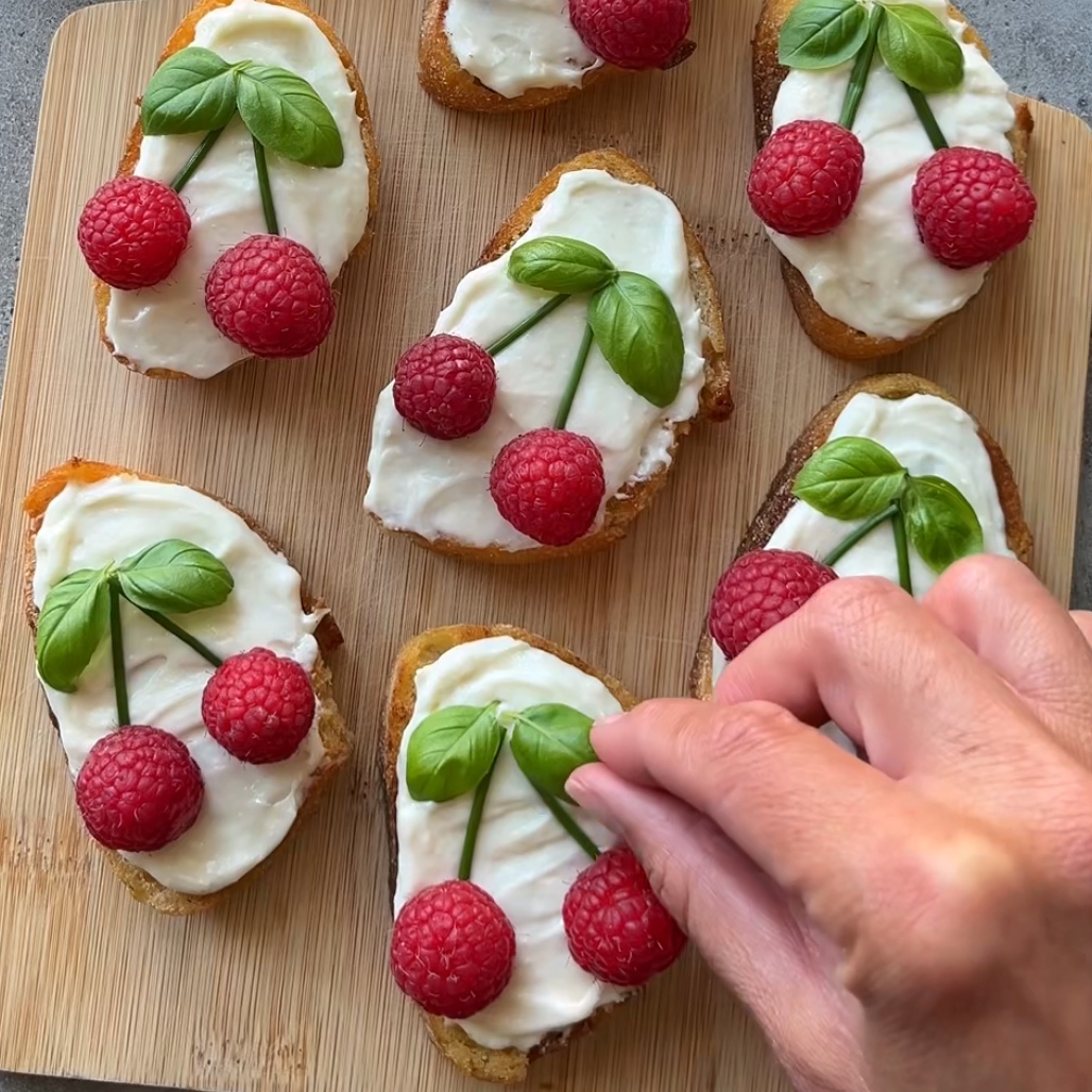 A hand decorates toast with cream cheese, fresh raspberries, and basil leaves arranged to resemble cherries on a wooden board.
