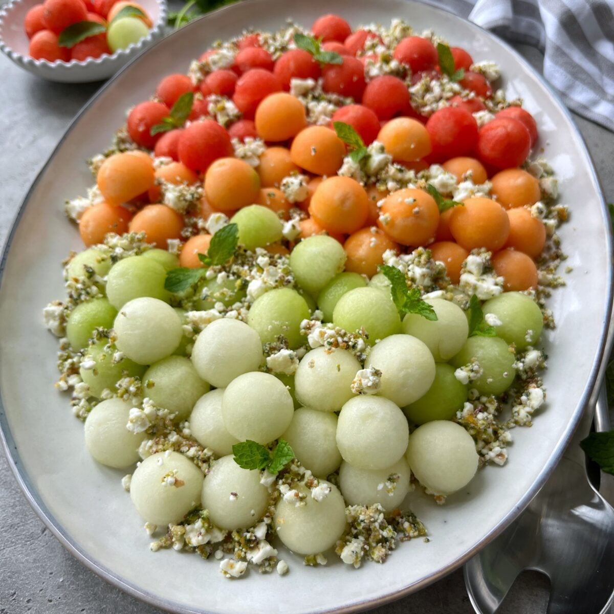 A vibrant Melon Salad platter of red, orange, and green melon balls, topped with crumbled cheese, herbs, and mint leaves, arranged neatly in rows on a white oval dish.