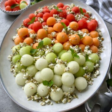 A vibrant Melon Salad platter of red, orange, and green melon balls, topped with crumbled cheese, herbs, and mint leaves, arranged neatly in rows on a white oval dish.