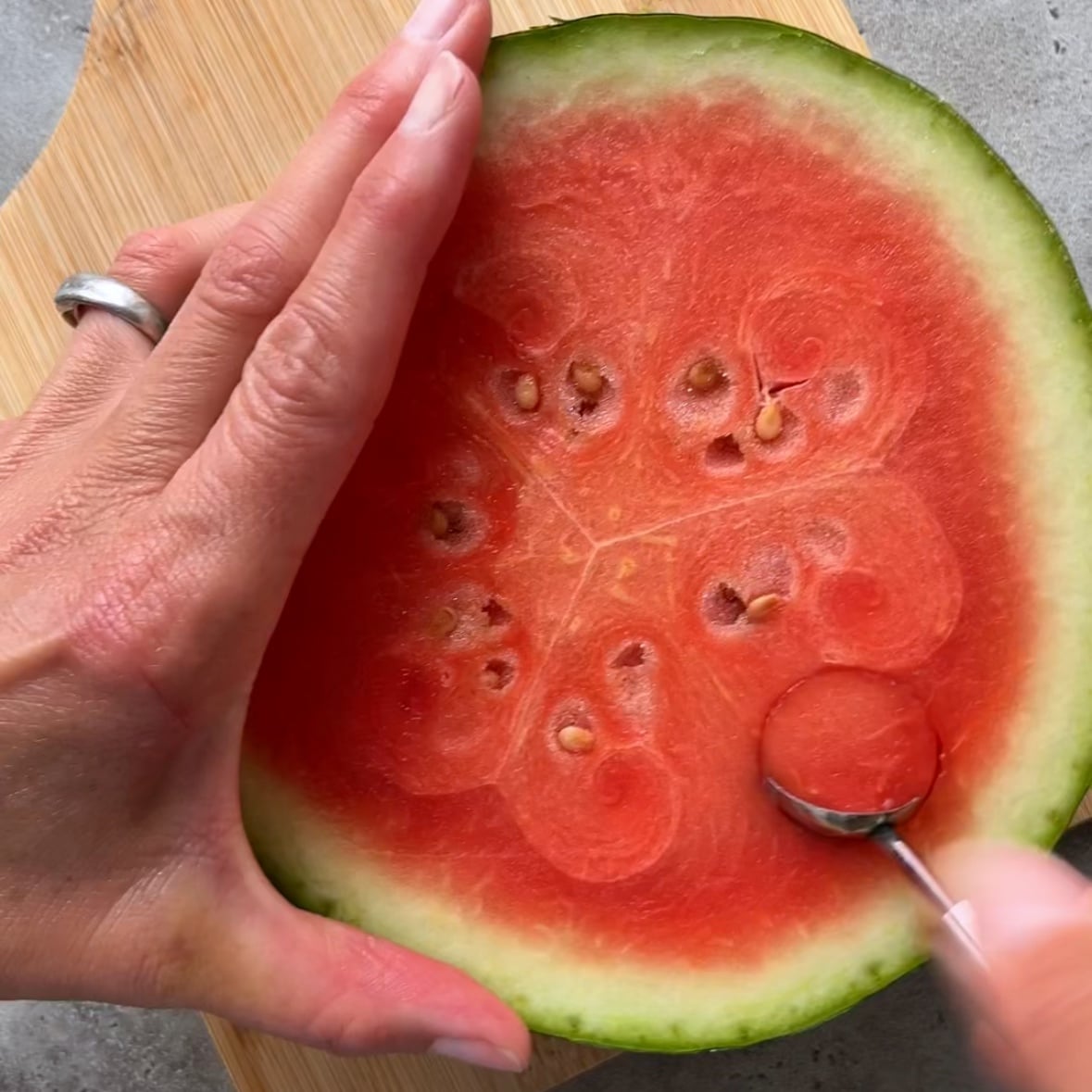 A hand holds half a watermelon on a cutting board while a small spoon scoops the red flesh.