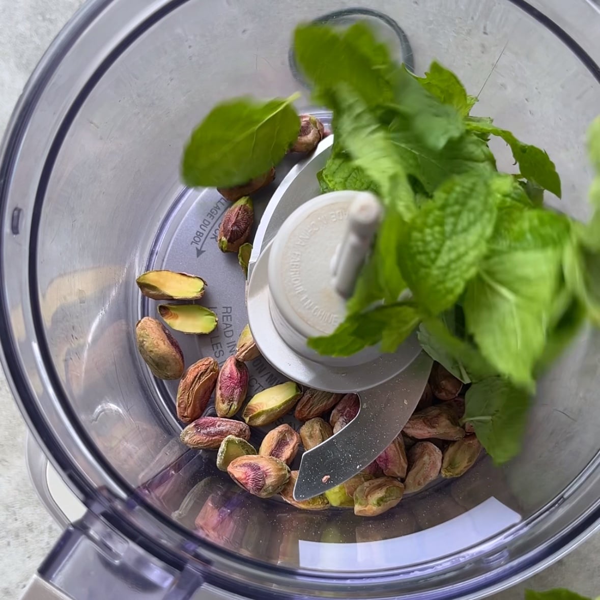 A food processor bowl with unshelled pistachios and fresh mint leaves inside, viewed from above.