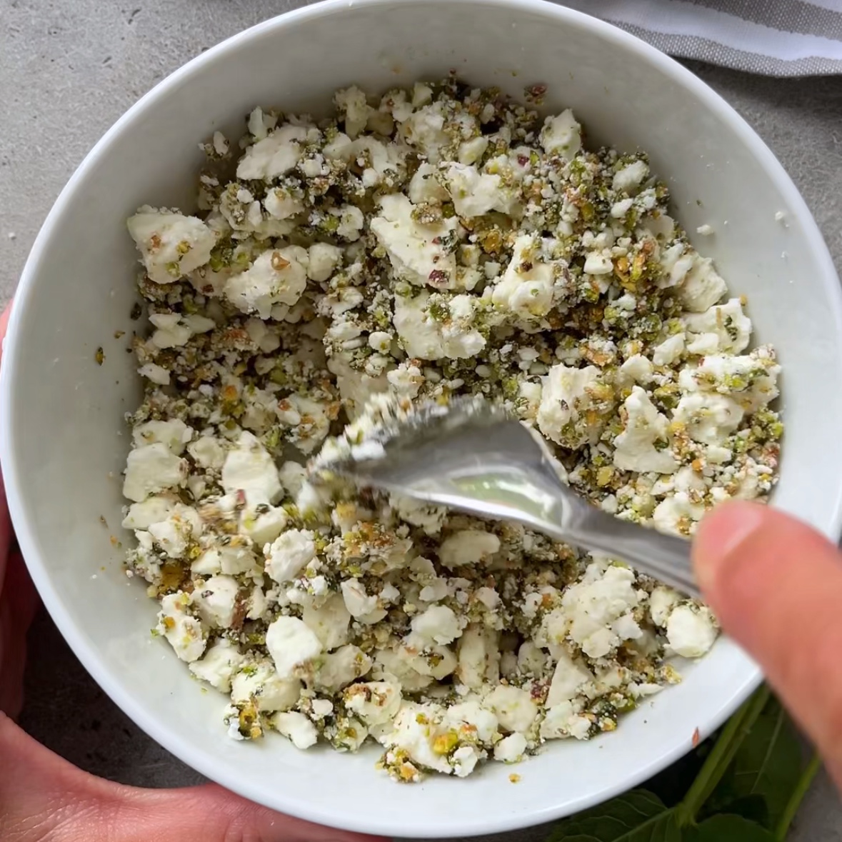 A hand holds a bowl of crumbled feta mixed with basil and nuts, creating the perfect topping for a refreshing Melon Salad.