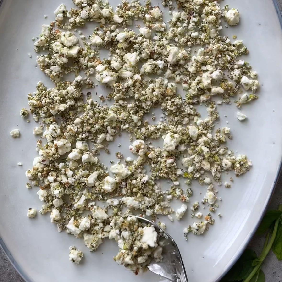 A plate with a scattered mixture of crumbled white cheese and herbs, with a spoon scooping some of the mixture at the edge of the plate.