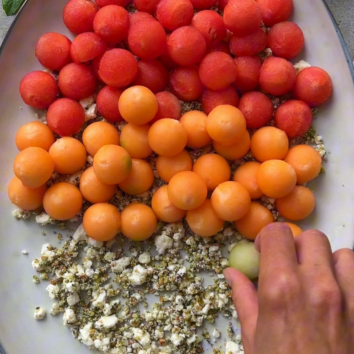 A hand arranges green melon balls on a platter with red watermelon balls and orange cantaloupe balls, atop a layer of crumbled cheese mixed with herbs.