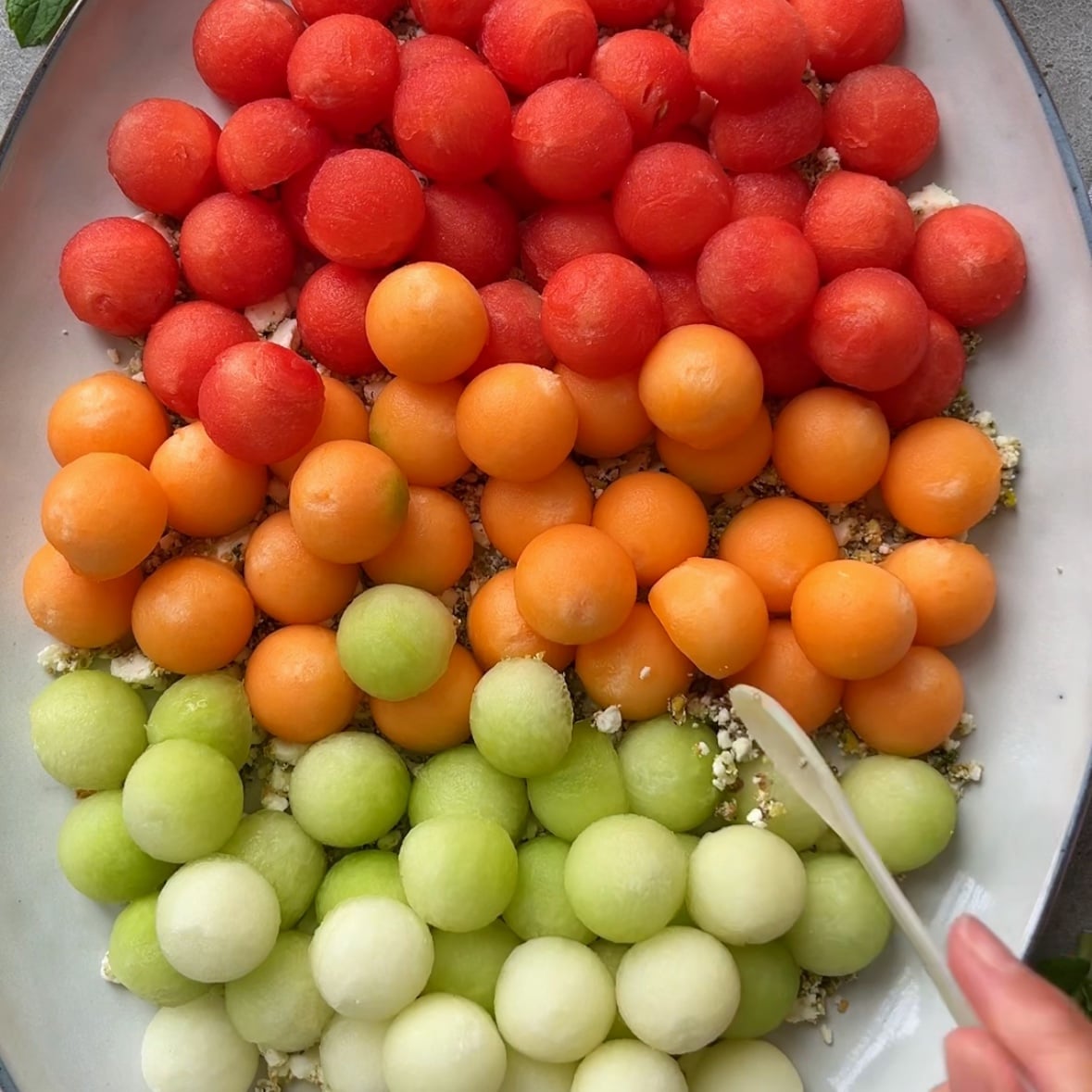 A vibrant Melon Salad platter with rows of watermelon, cantaloupe, and honeydew melon balls, arranged by color, with a hand holding a spoon and topping a crumbly feta mixture at the bottom right.