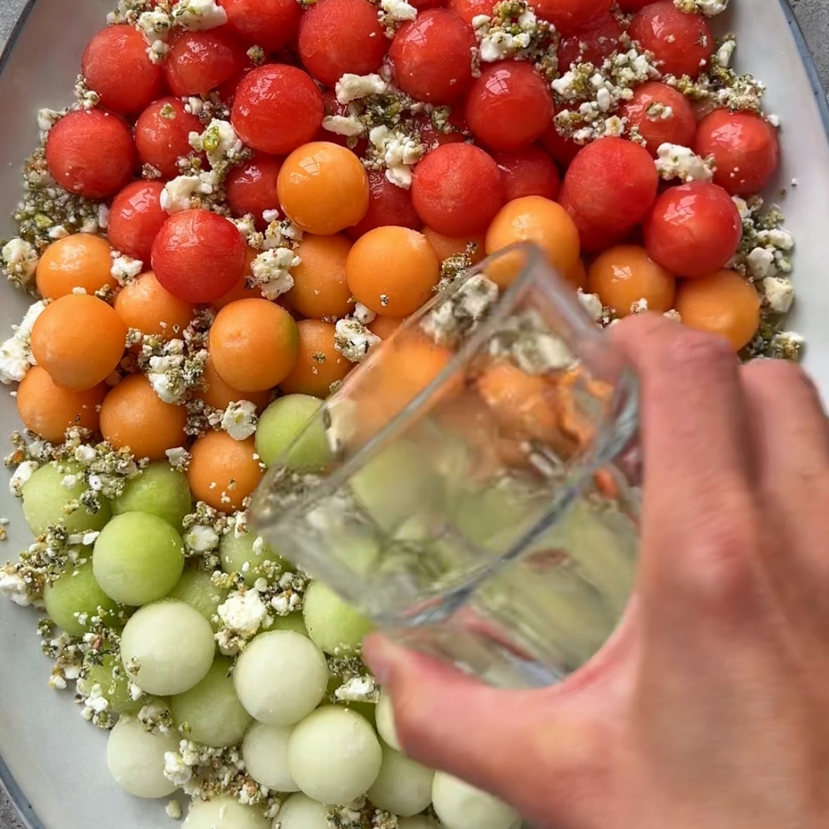 A hand pours dressing from a glass over a platter of red, orange, and green melon balls mixed with crumbled cheese and herbs.