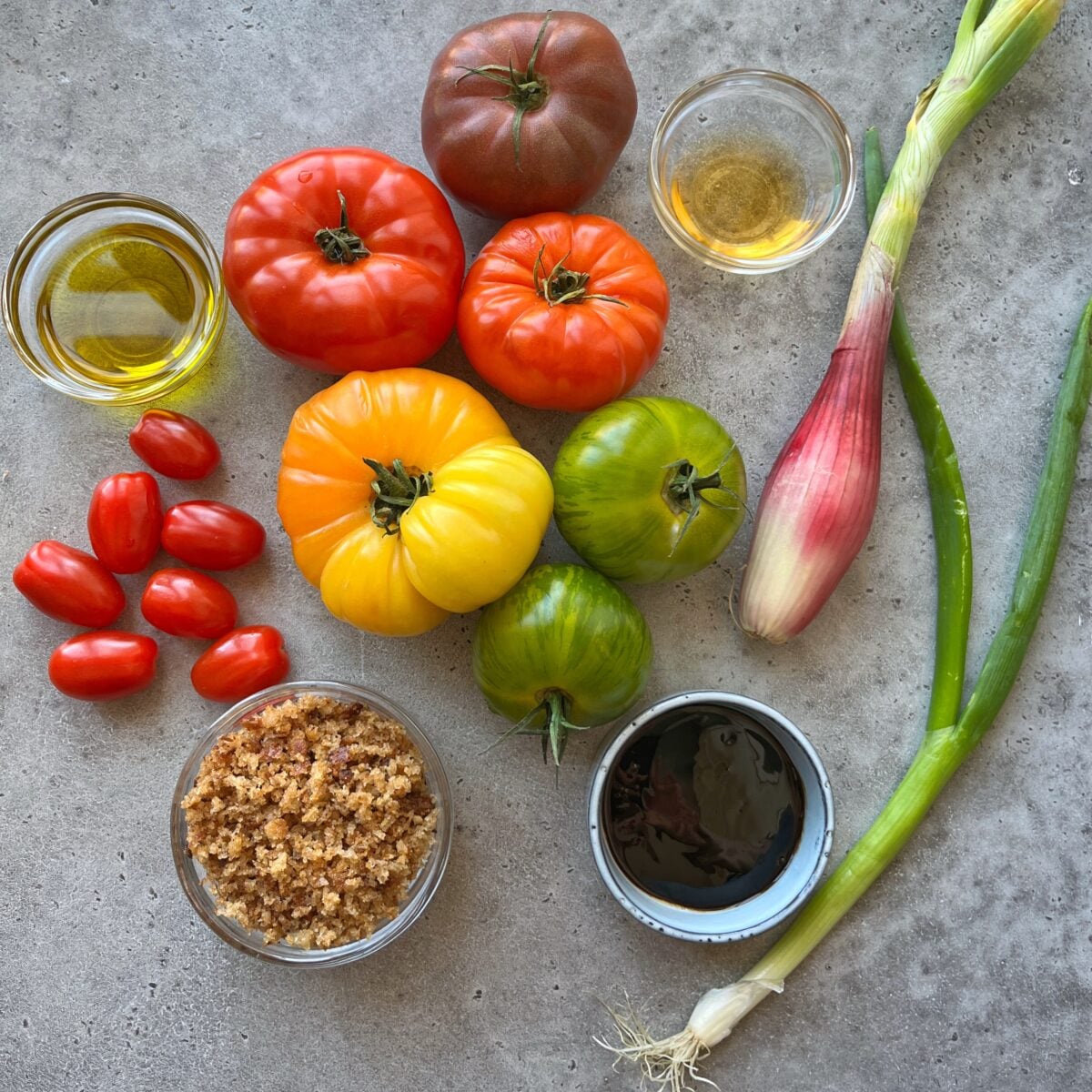 Assorted heirloom tomatoes, grape tomatoes, shallots, green onions, olive oil, vinegar, balsamic glaze, and breadcrumbs arranged on a gray surface.