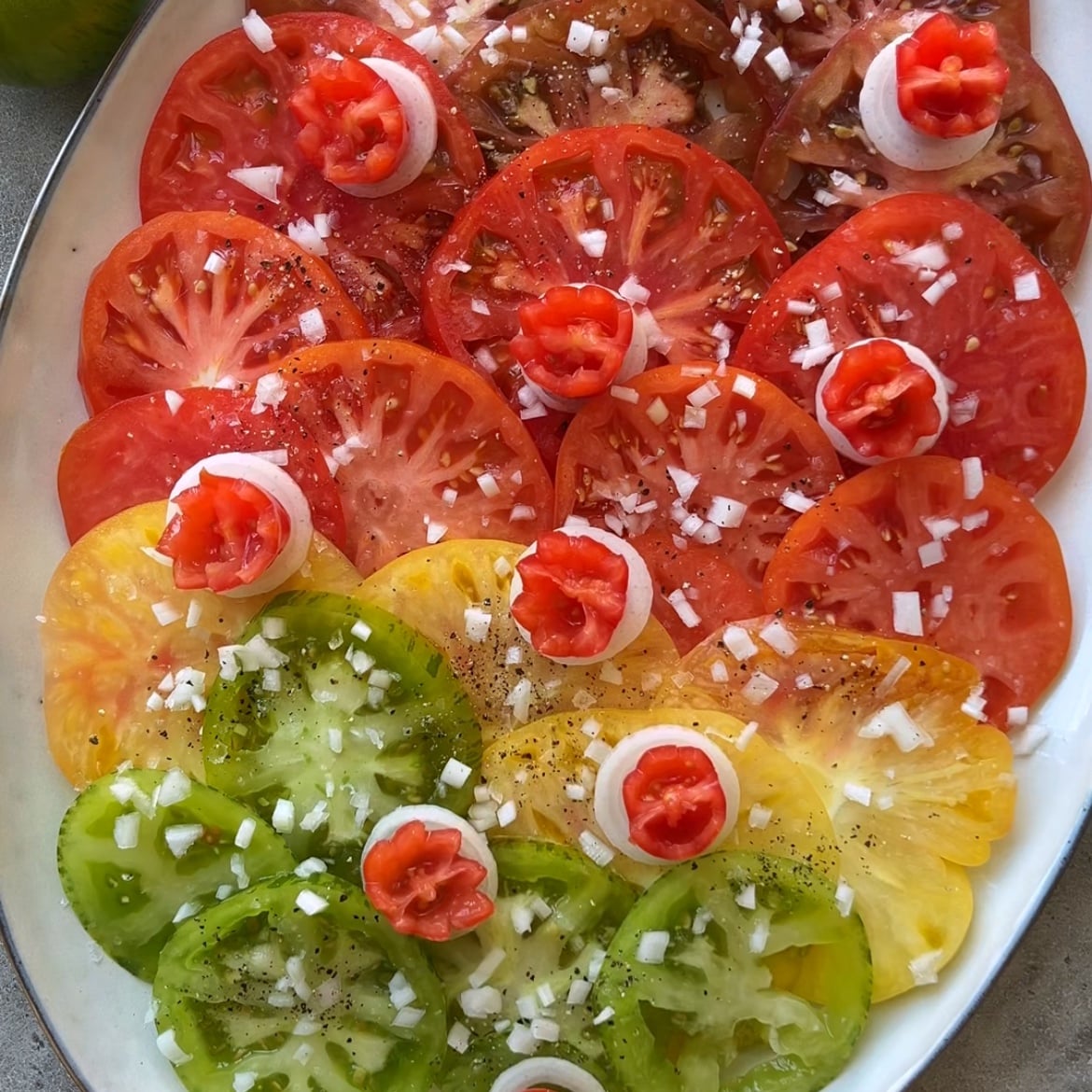 Heirloom tomato salad featuring sliced red, yellow, and green tomatoes arranged on a platter, garnished with diced onions, black pepper, and small tomato and onion rosettes.