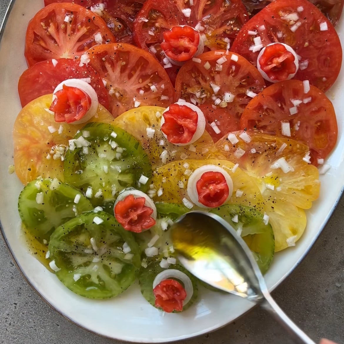 A vibrant heirloom tomato salad platter features sliced red, yellow, and green tomatoes topped with chopped onions, mini tomatoes, black pepper, and a drizzle of vinaigrette