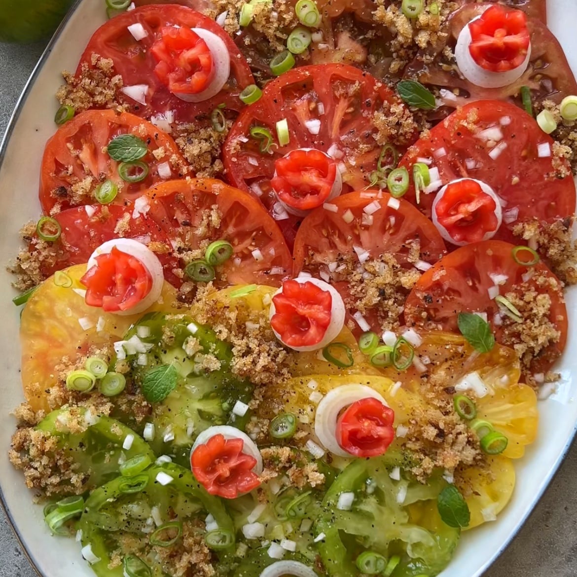 A vibrant heirloom tomato salad features sliced red, yellow, and green tomatoes on a platter, garnished with breadcrumbs, scallions, fresh herbs, and tomato flowers.