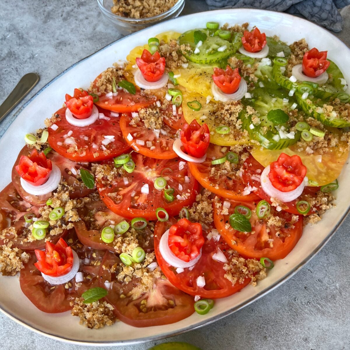Heirloom Tomato Salad platter with sliced red, green, and yellow tomatoes, topped with onion rings, tomato garnishes, chopped green onions, and golden baked breadcrumbs.