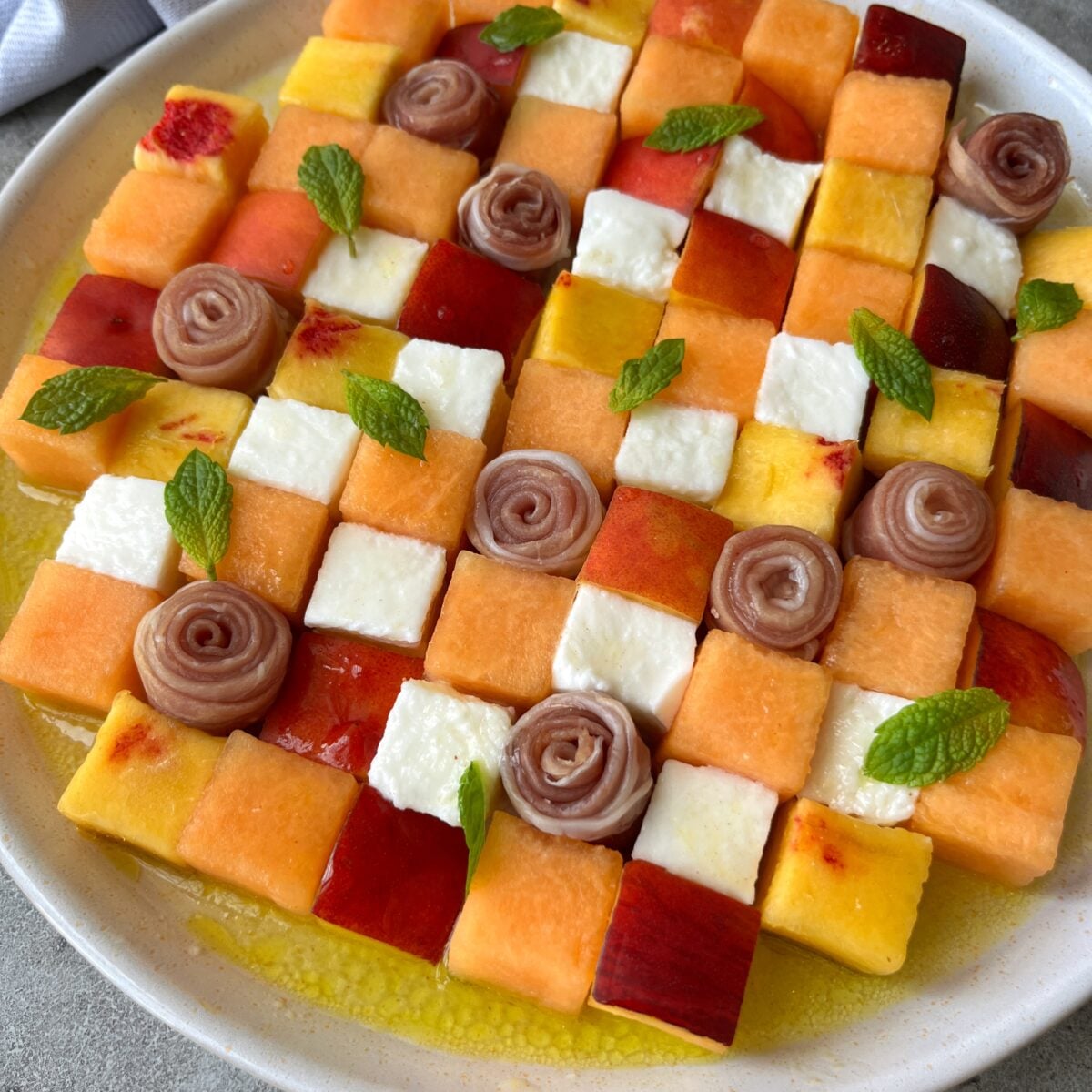 A plate arranged with a checkerboard pattern of melon, prosciutto rosettes, nectarine, and mozzarella cheese cubes.