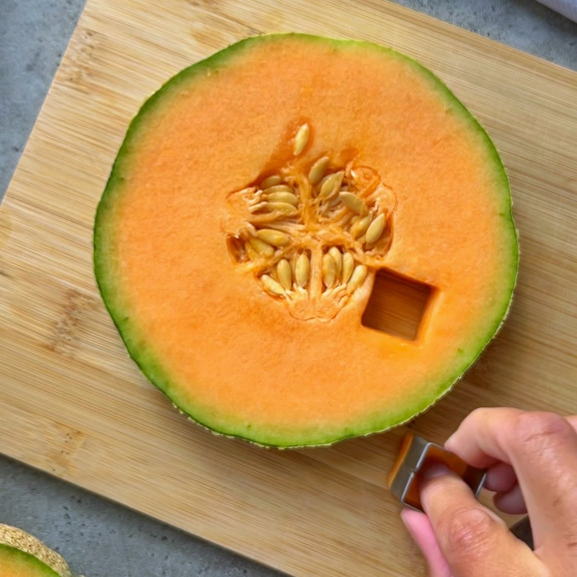 A hand uses a small square cutter to remove a cube from a halved cantaloupe on a wooden cutting board.