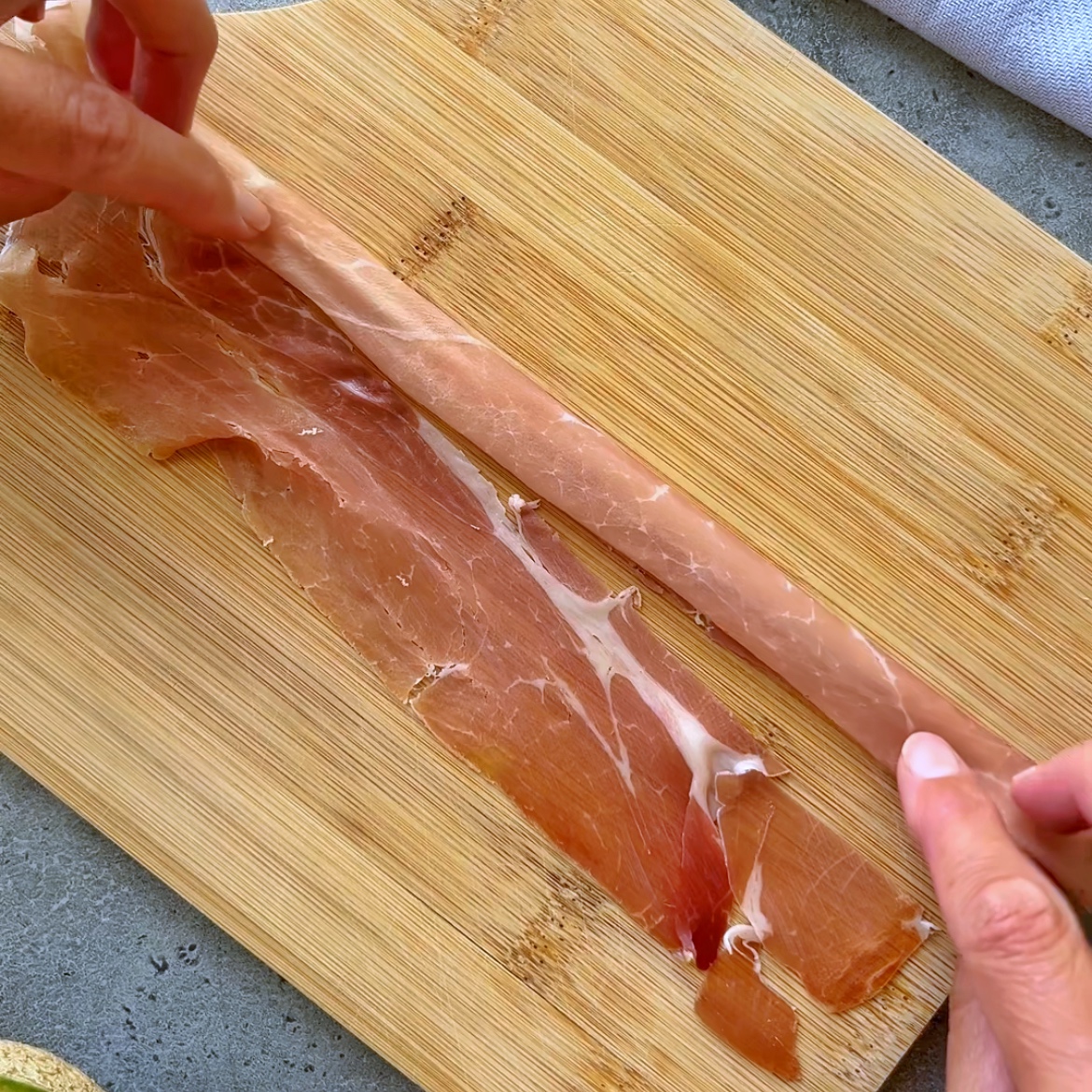 Two hands roll a thin slice of  prosciutto, on a wooden cutting board.