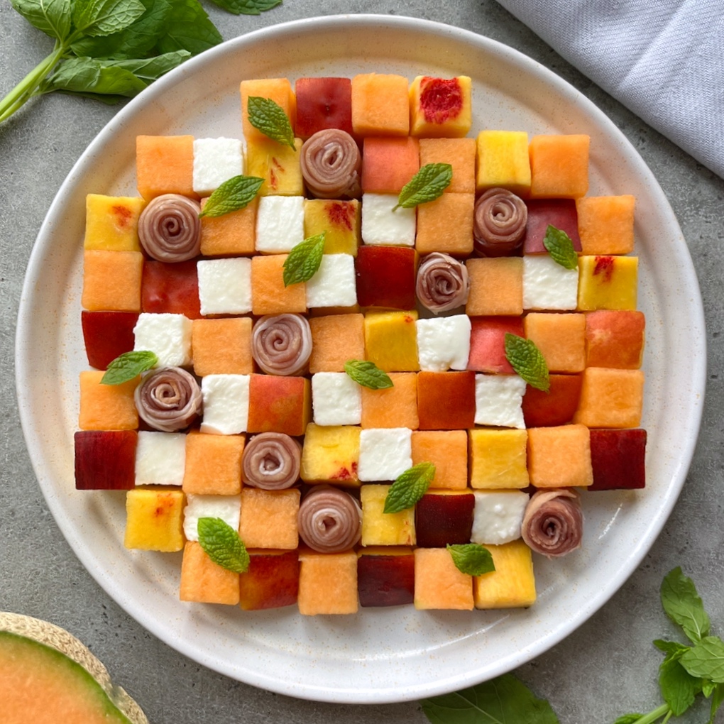 A plate arranged with colorful fruit, cheese cubes, and Melon Prosciutto Salad in a grid pattern, garnished with small mint leaves.