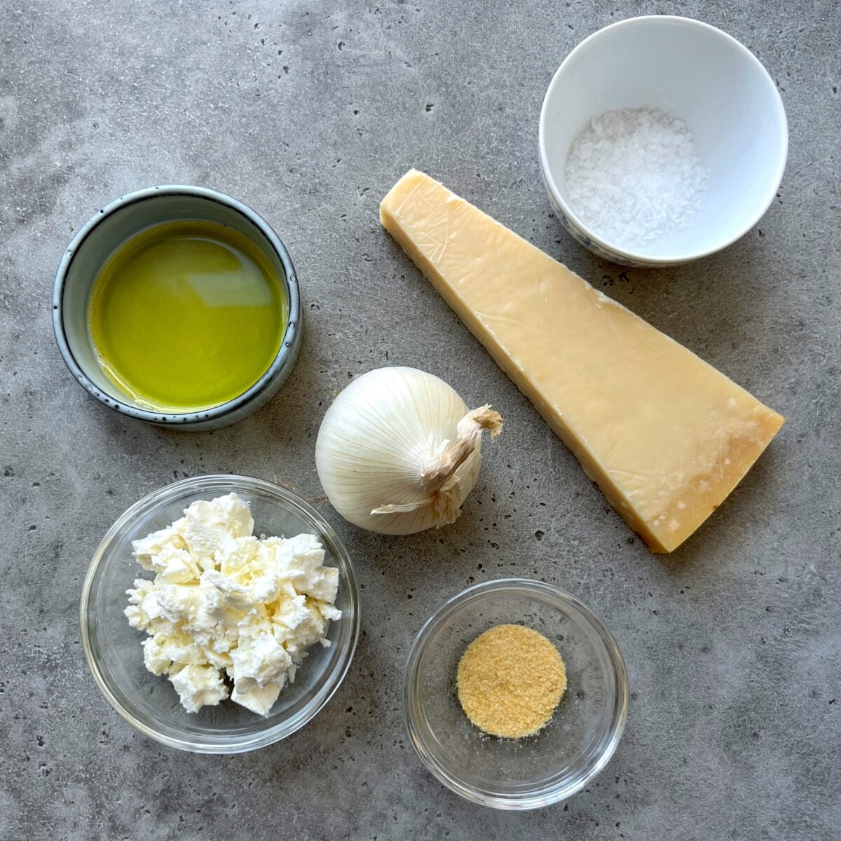 Ingredients on a gray surface: a wedge of Parmesan, a white onion, olive oil, salt, crumbled cheese, and seasoning.