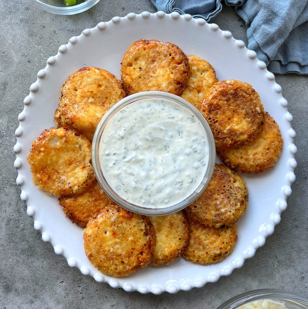 A white plate with crispy, round Parmesan chips with onion arranged around a bowl of creamy dip on a gray surface.