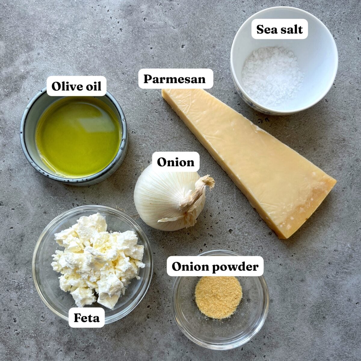 Various labeled ingredients on a gray surface, including olive oil, sea salt, parmesan cheese, a whole onion, crumbled feta, and onion powder in small bowls.
