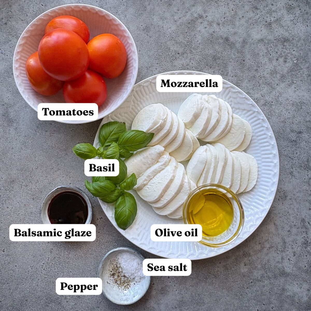 A plate with sliced mozzarella, basil, a bowl of tomatoes, olive oil, balsamic glaze, sea salt, and pepper on a gray surface.