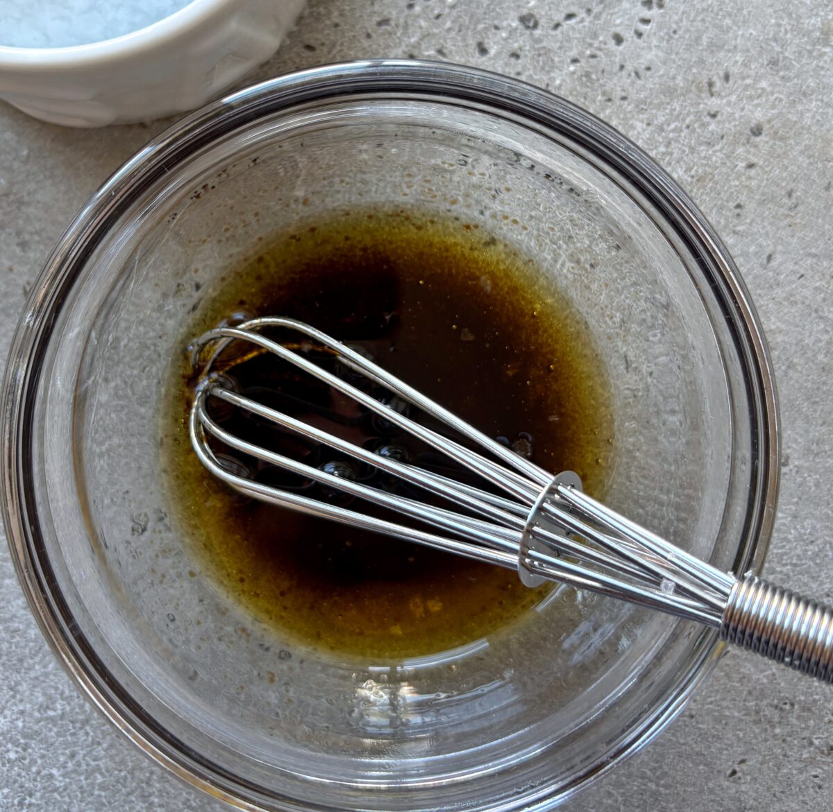 A metal whisk rests in a glass bowl containing a balsamic glaze and olive oil dressing. A white bowl with coarse salt is partially visible in the top left corner, sitting on a gray countertop.