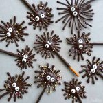 Chocolate spider treats with candy eyes and sprinkles arranged on a grey surface. These playful bites have thin, spiky legs made by drizzling chocolate over stick-shaped bases—a fun addition to your favorite food recipes.