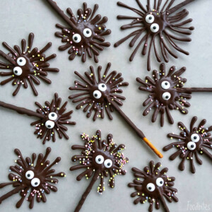 Chocolate spider treats with candy eyes and sprinkles arranged on a grey surface. These playful bites have thin, spiky legs made by drizzling chocolate over stick-shaped bases—a fun addition to your favorite food recipes.