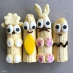 Four banana halves decorated as cute animals and characters with candy and chocolate, featuring big eyes and playful details like ears, paws, and whiskers—adorable bites perfect for fun food recipes or healthy treats on a gray background.
