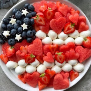 A patriotic platter inspired by food recipes, arranged like the American flag with blueberries and blackberries topped with white star-shaped fruit, rows of mozzarella balls, watermelon hearts, and strawberry slices—perfect for festive bites.