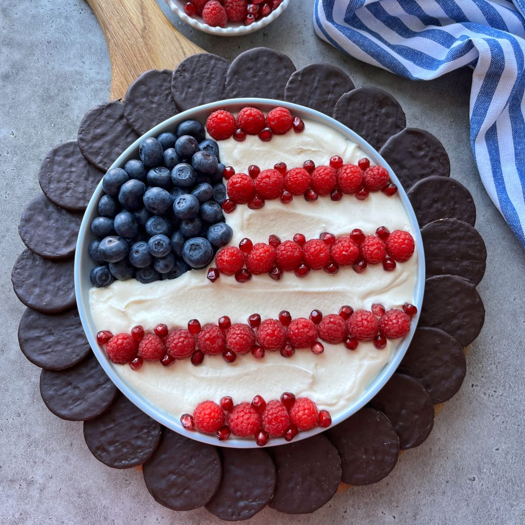 A round 4th of July dessert dip platter decorated like the American flag with blueberries, raspberries, and pomegranate seeds on a creamy base, surrounded by chocolate cookies. A striped blue and white cloth is nearby.