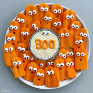 A plate of carrot slices shaped like ghosts with candy eyes surrounds a bowl of dip. This fun dish features the word “BOO” spelled out with carrot letters in the center.
